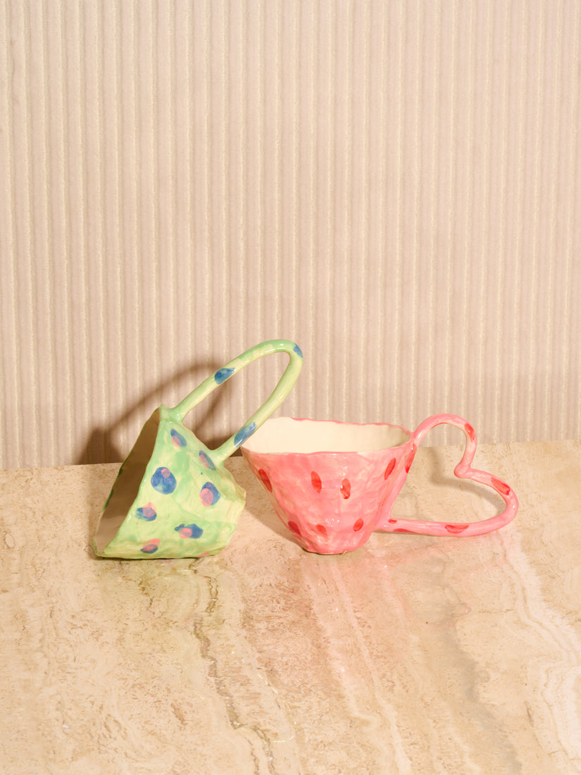 Green Mug with Peas leans on its side with its handle resting on the lip of the Pink Heart Mug.