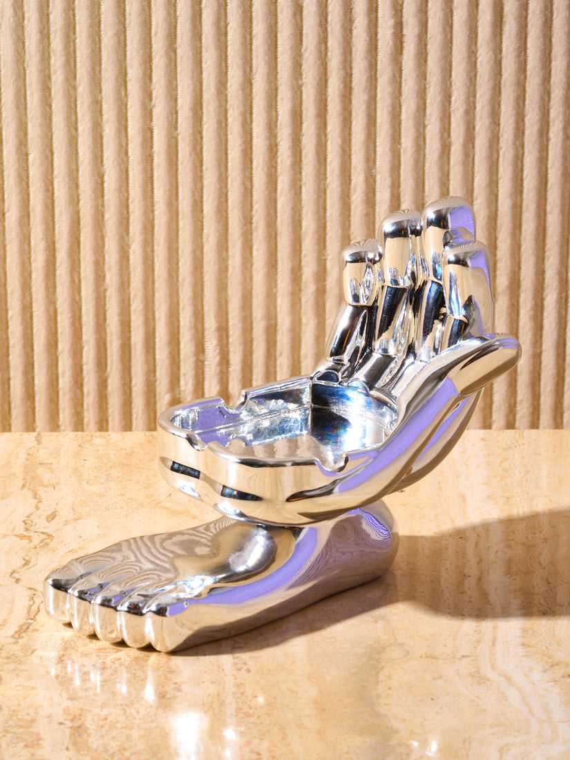 Hand and foot chrome ashtray. Hand is shown palm up with notches to hold cigarette or joint. Connected to the palm on the bottom half shows a foot.This object is displayed on a cream marble table.