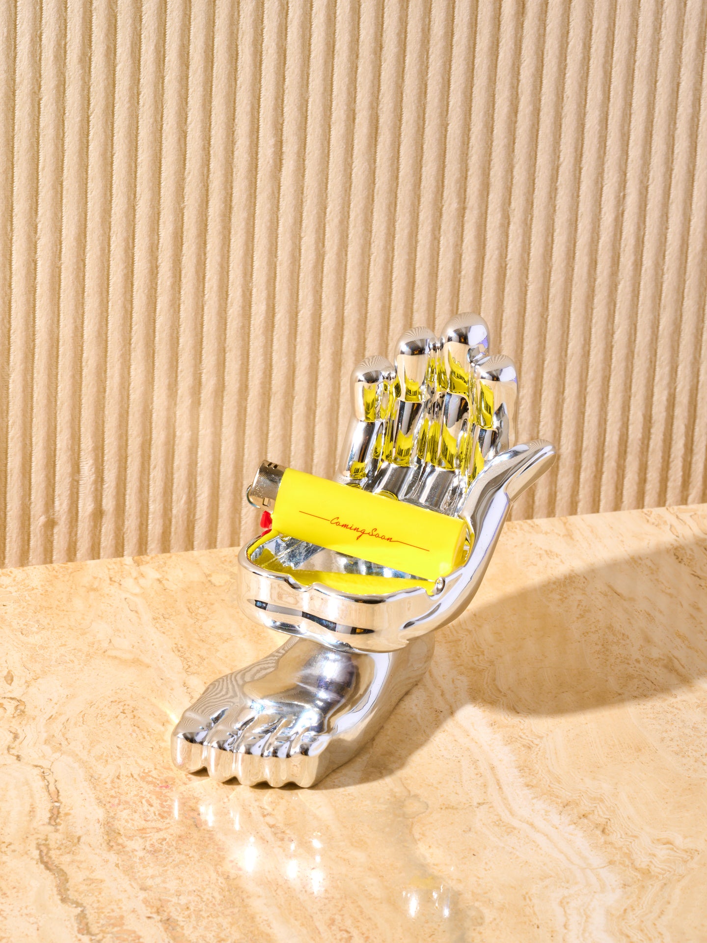 Hand and foot chrome ashtray displayed on a cream marble table with a yellow lighter in the notches reading "coming soon".