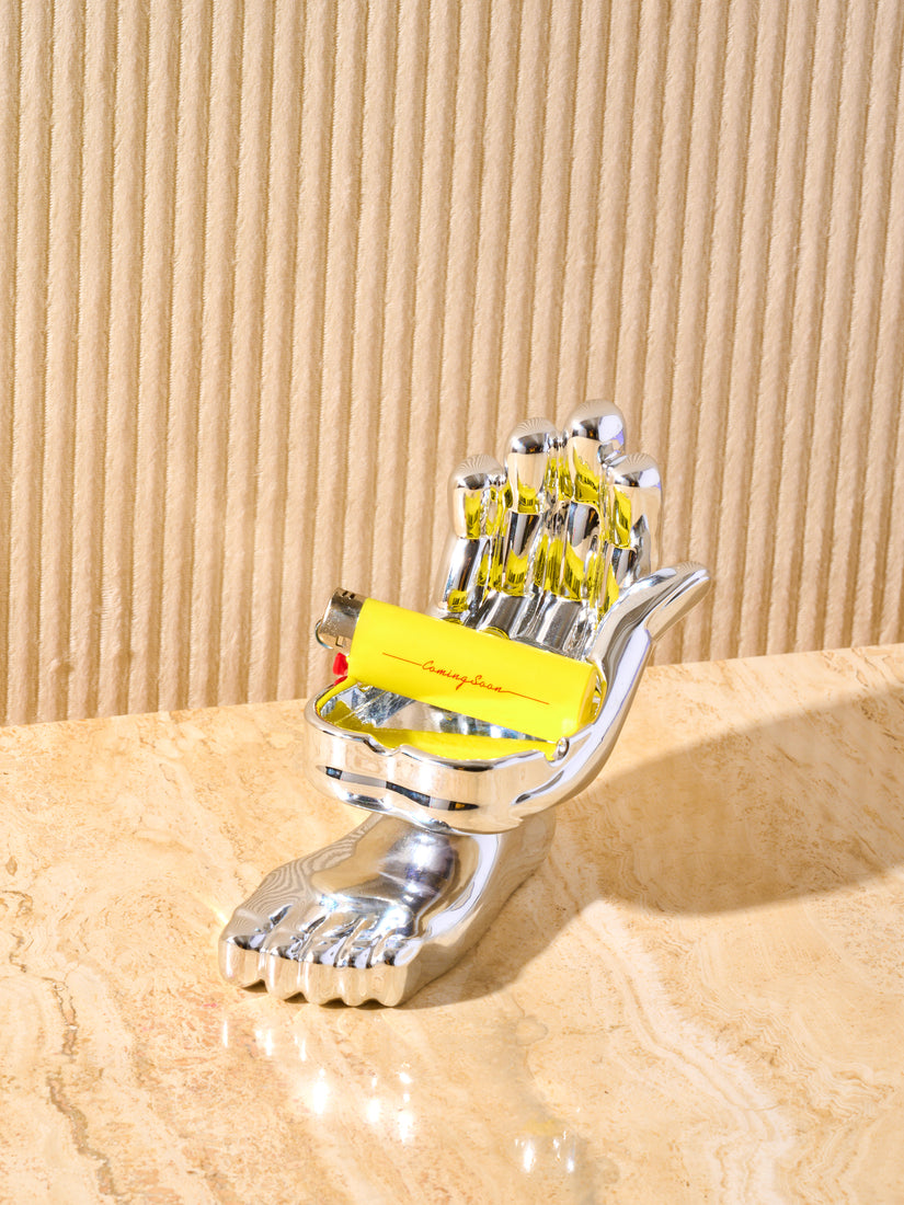 Hand and foot chrome ashtray displayed on a cream marble table with a yellow lighter in the notches reading "coming soon". 