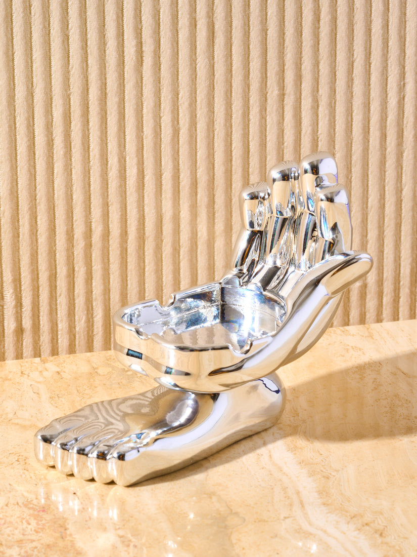 Hand and foot chrome ashtray. Hand is shown palm up with notches to hold cigarette or joint. Connected to the palm on the bottom half shows a foot.This object is displayed on a cream marble table.