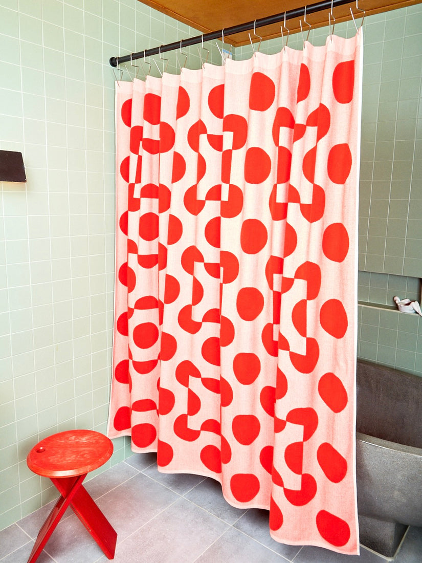 A red and salmon colored terry cloth shower curtain with circle and square shapes by Dusen Dusen hung in a sage green tile bathroom.  A red stool sits to the left of the curtain. 