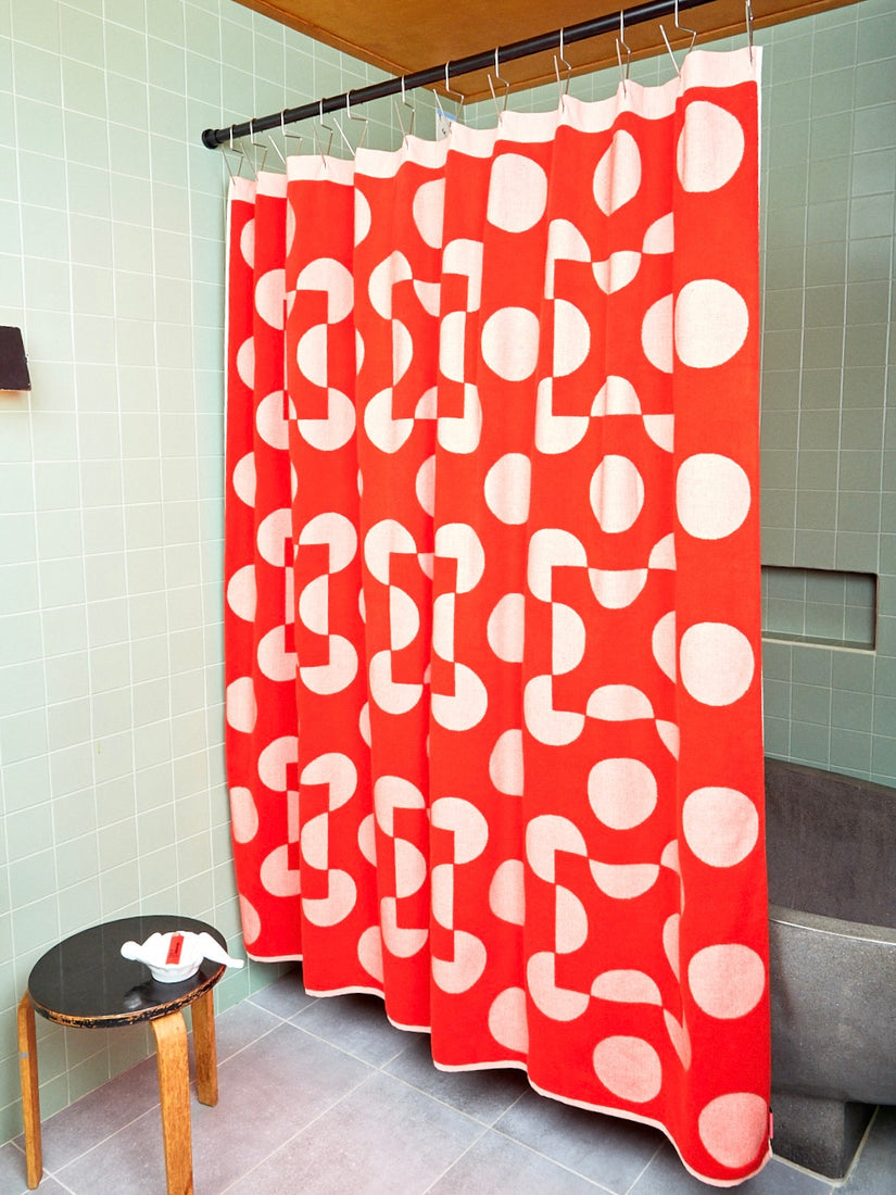 Red and Salmon shower curtain is shown in reverse in the sage green tiled bathroom. A black stool with a white bathing lady and red lighter sit to the left of the curtain. 