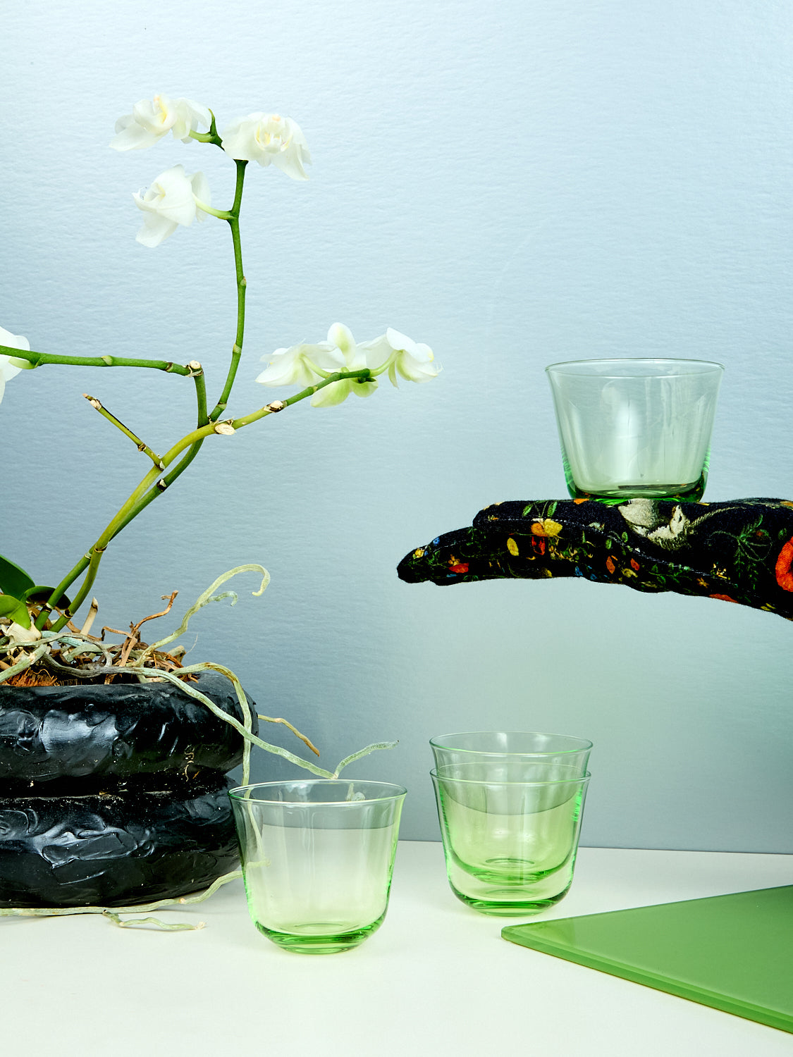 Green glass wine glasses are displayed with a hand reaching for one of them, alongside a floral arrangement.