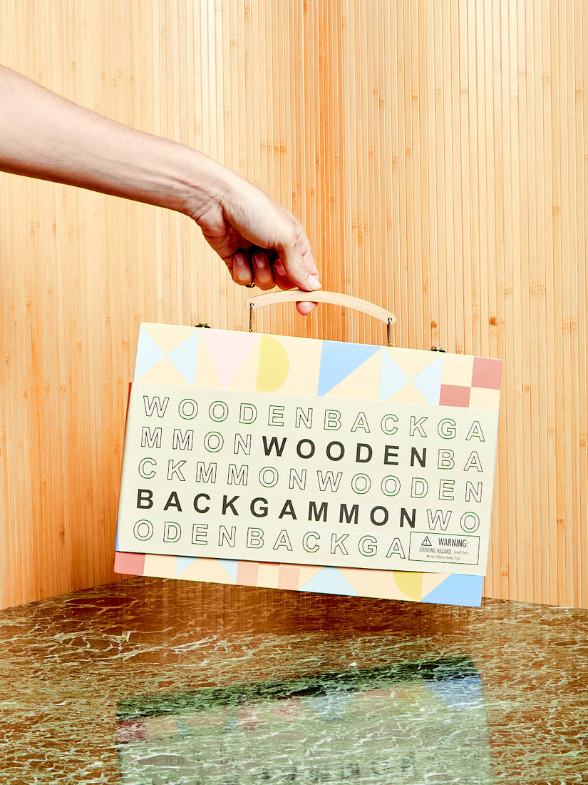 A hand holds Sunnylife's Wooden Backgammon game up by its handle. The game is in its branded graphic packaging sleeve.