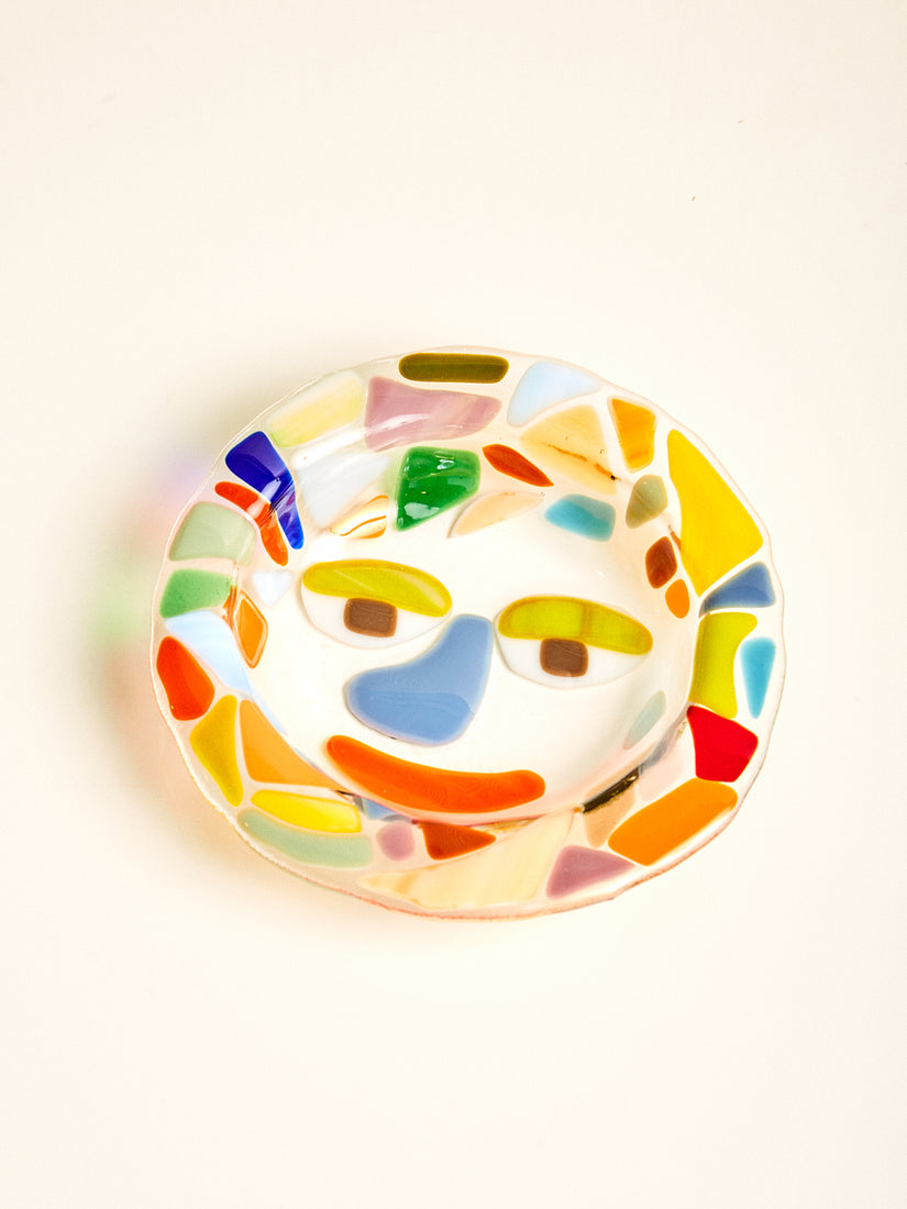 A clear glass bowl with a face design surrounded by a multicolor mosaic design.