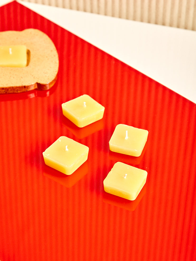 Four butter pat shaped beeswax tealight candles sit on a red glossy surface. In the background the toast candle holder sits slightly out of frame.