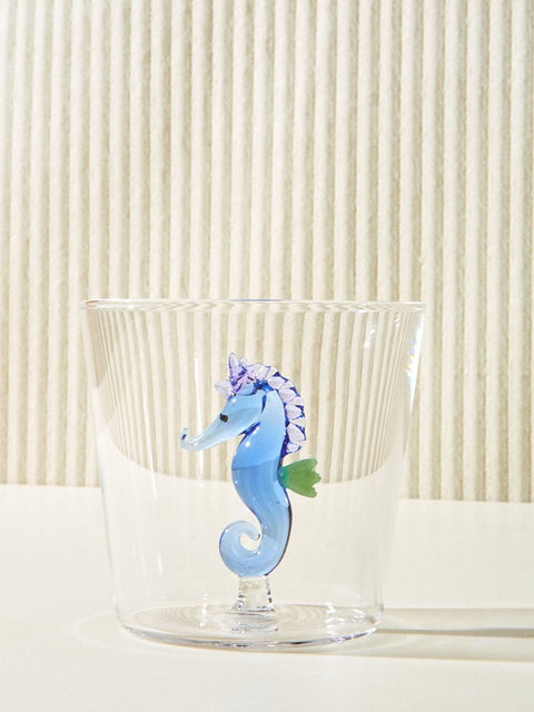 A small cup with a blue seahorse made of glass inside it sits on a cream table.