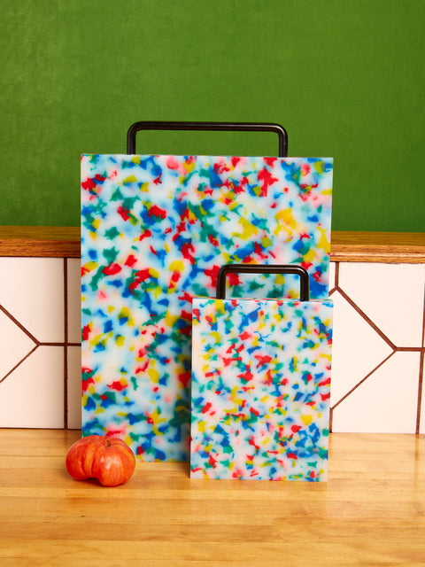 A multicolored plastic chopping block with a metal handle, displayed on a wooden surface with a green tiled wall in the background.
