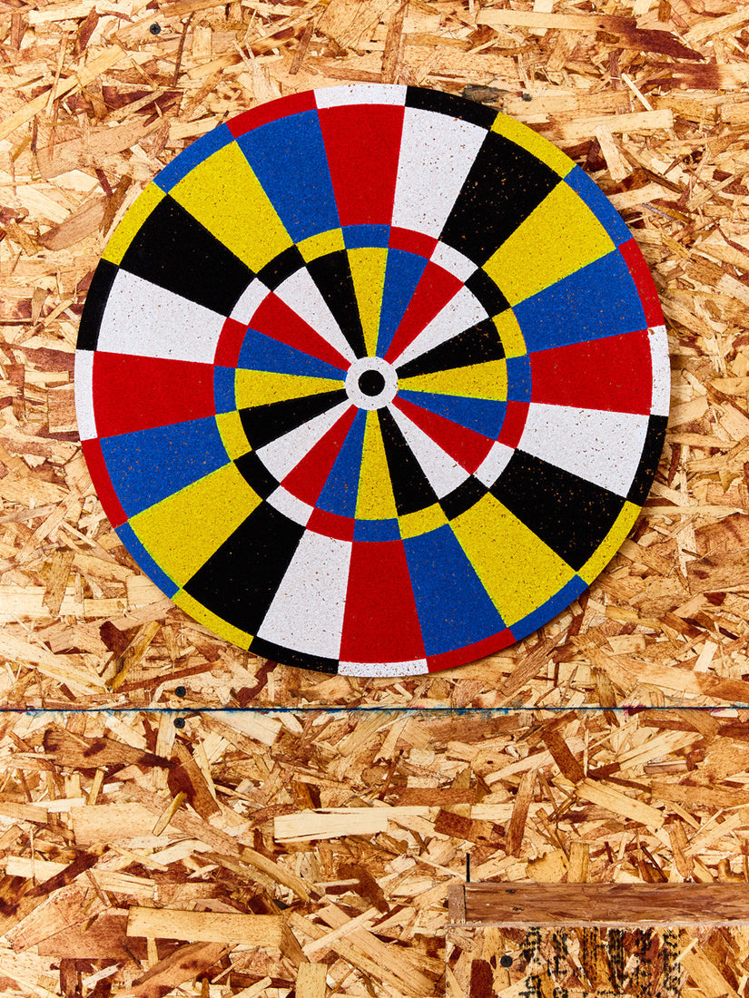 Red, yellow, blue, black, and white painted cork Dart Board by Fredericks and Mae on a plywood wall.