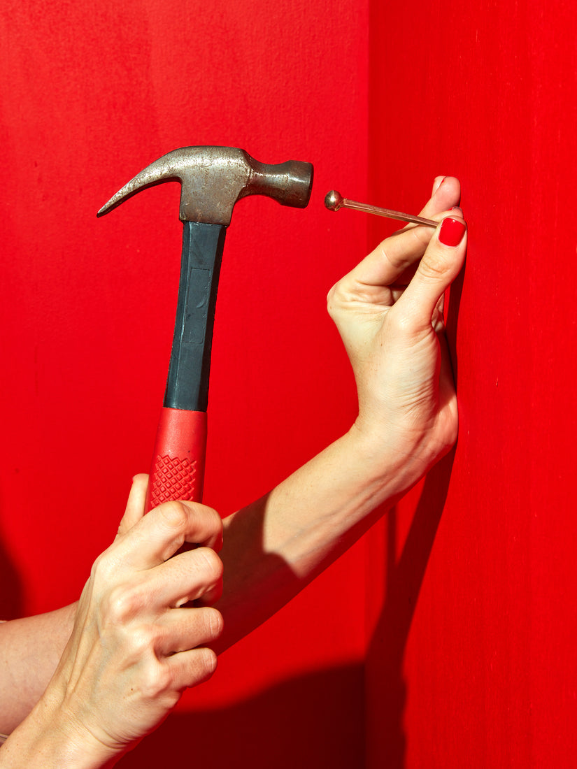 On a vibrant red background are two hands, one holding a Coming Soon Hester the Nail against the wall, and another yielding a hammer.