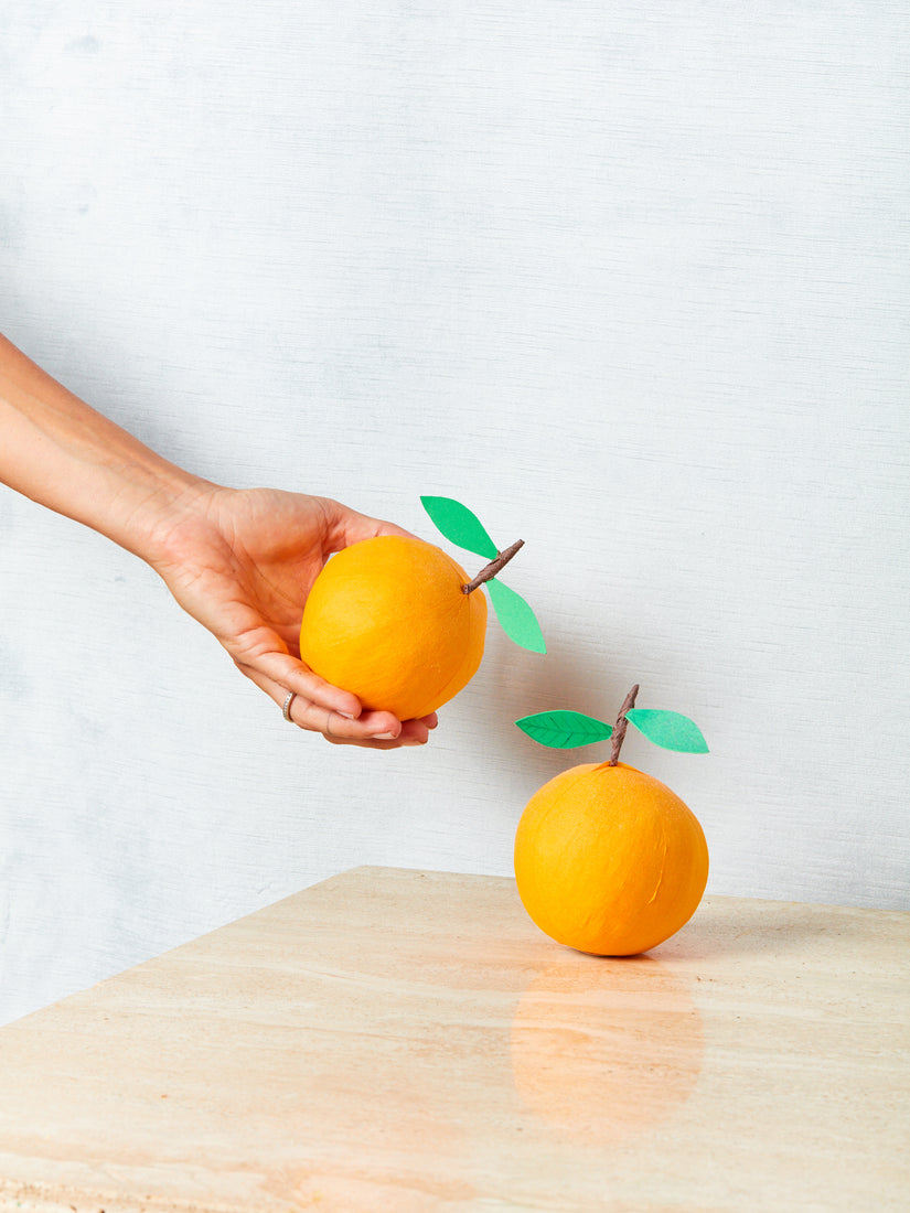 A hand holds up an Orange Suprise Ball next to another that rests on a table top.