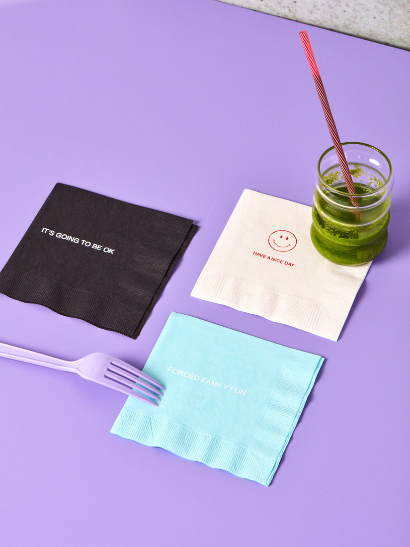 Three cocktail napkins in different colors with different prints: IT'S GOING TO BE OKAY, HAVE A NICE DAY, FORCED FAMILY FUN