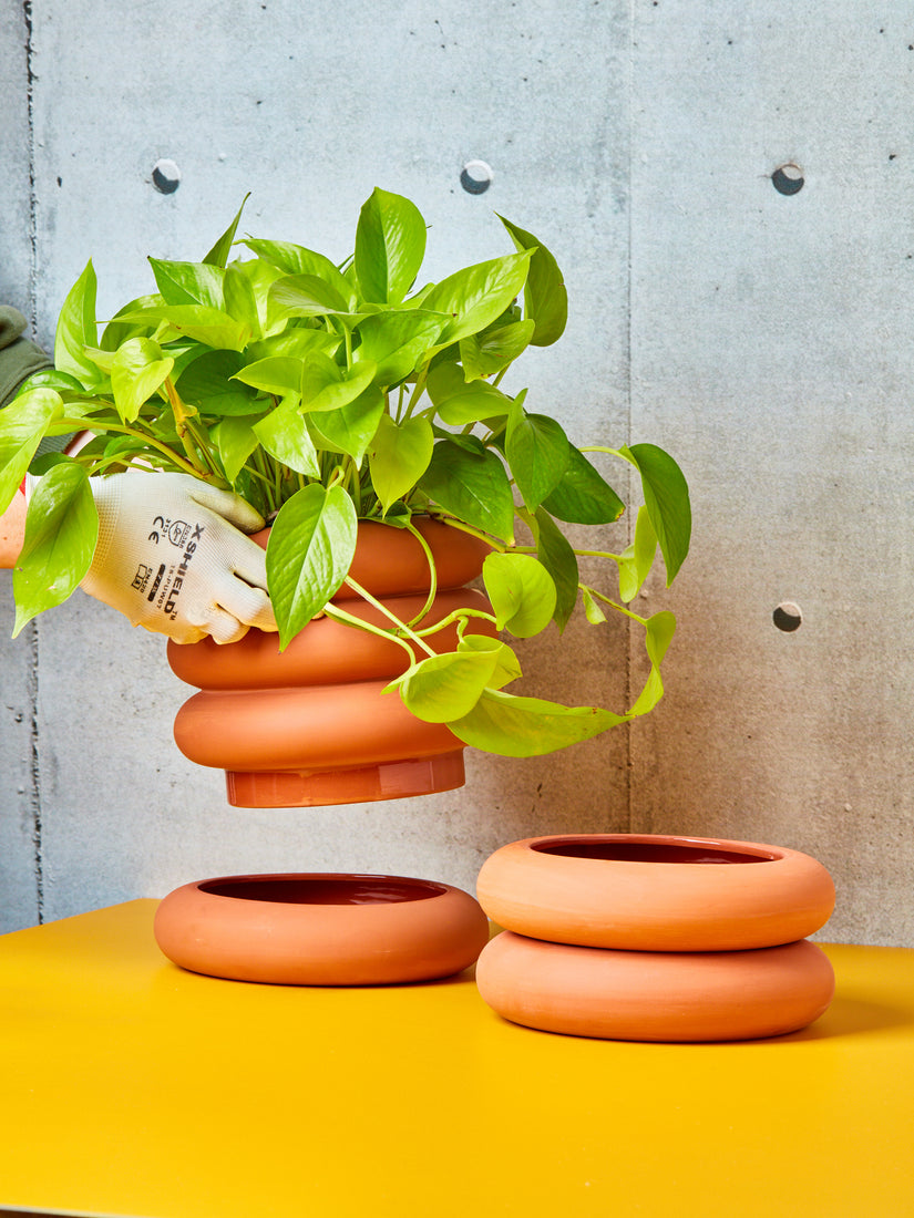 Hands lift up the tall terracotta stacking planter from its saucer. A pothos plant is potted inside the planter, and the short version sits right.