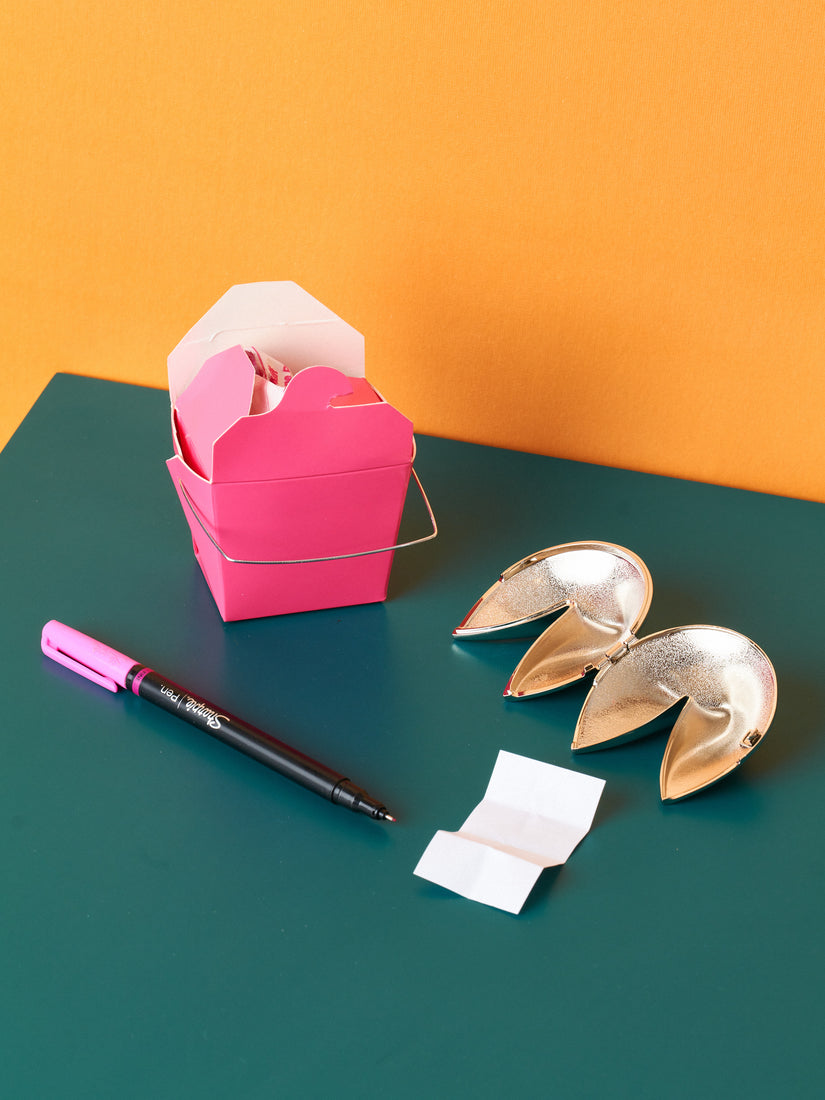 A pink take out box, pink sharpie, blank slip of paper, and open chrome fortune cookie.