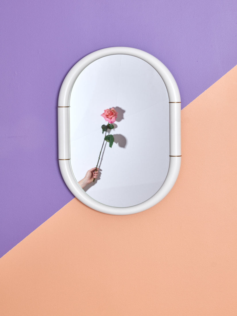Ceramic Mirror in white by Entler Studio hung on a lavender and peach wall. A hand holding a pink rose can be seen in the reflection.