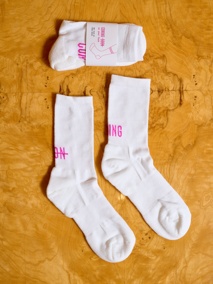 Two pairs of white Coming Soon socks, one in its packaging.