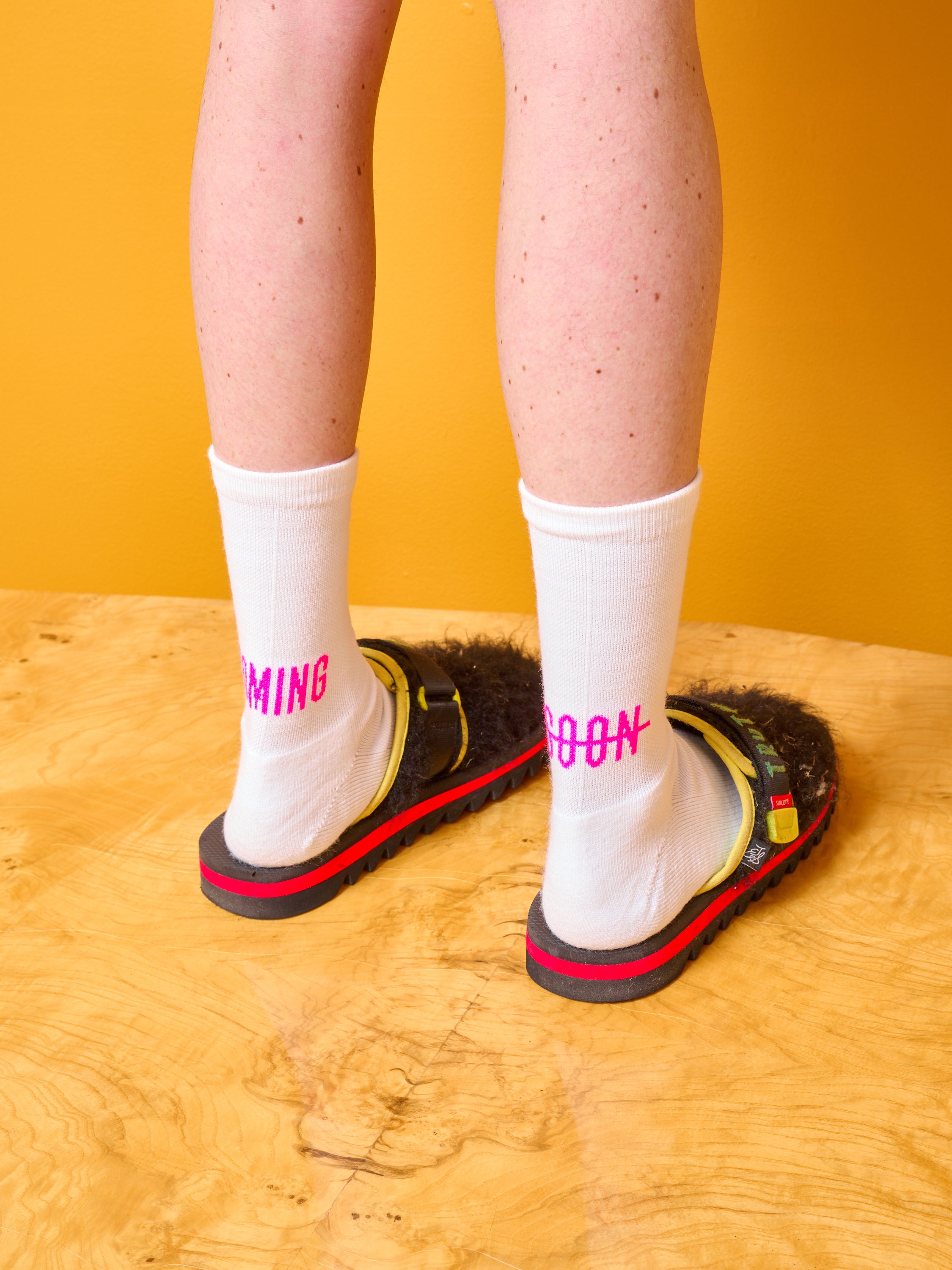 Someone wearing white Coming Soon socks with pink logo.