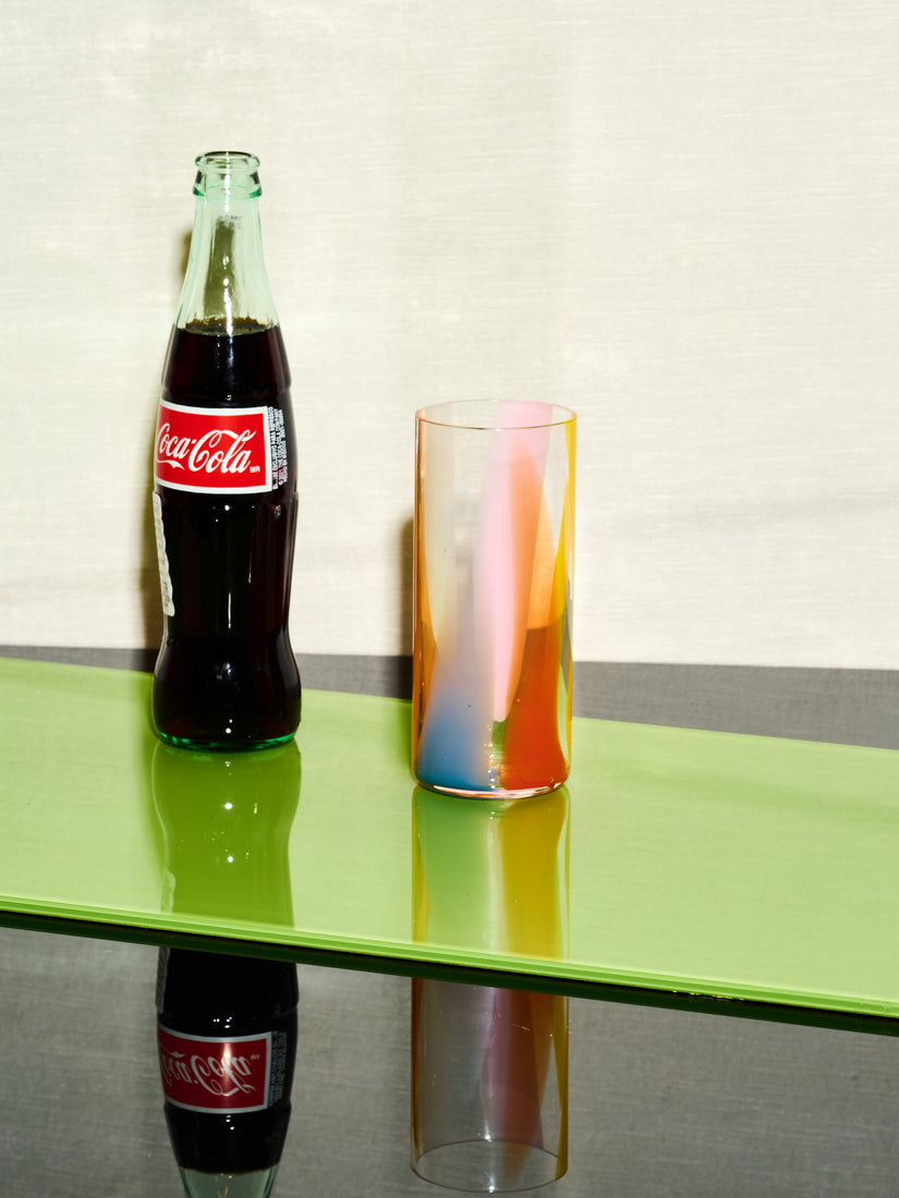 A glass bottle of coke sits left of a Rainbow Splash Highball by Bow Glass.