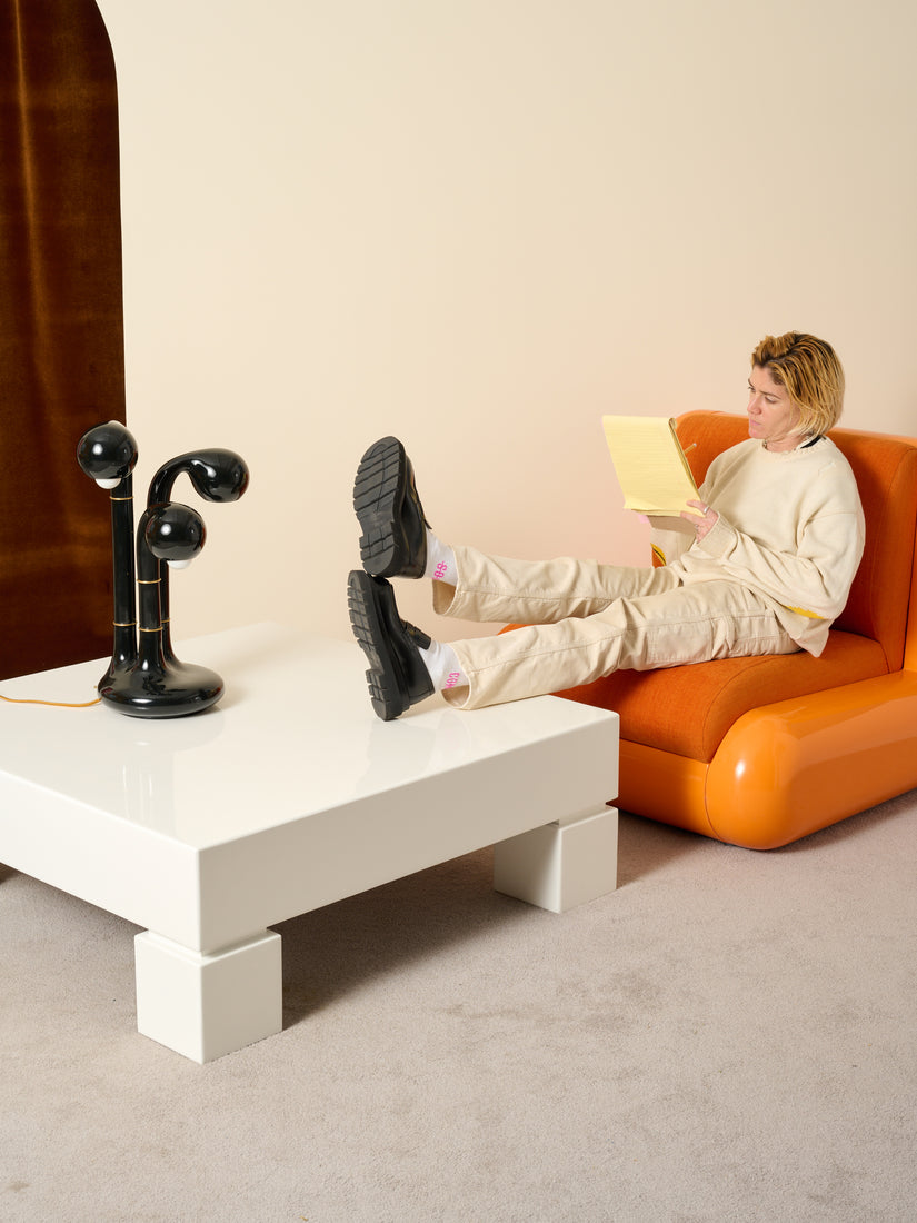 A person writes in a legal pad while sitting in an orange Uma T4 Chair with their heels propped onto a large white coffee table. Atop the coffee table sits a three globe table lamp by Entler in glossy black glaze.