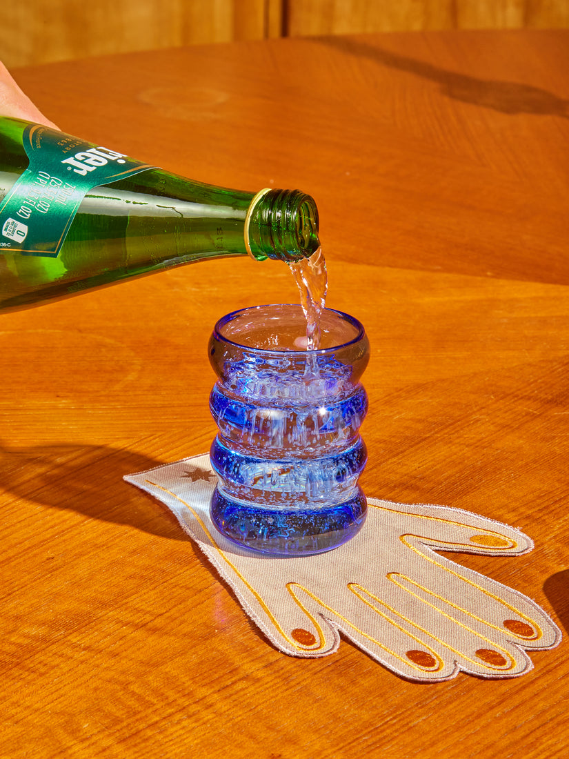 A glass bottle of Perrier being poured into a blue Ripple Glass resting on a hand cocktail napkin.