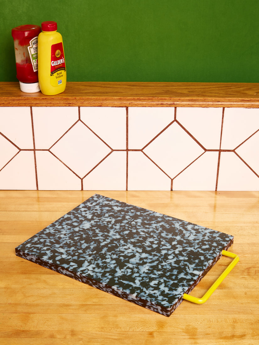 A black and white chopping block by fredericks and mae with a yellow handle sits on a kitchen counter. On the ledge of the kitchen counter are ketchup and mustard bottles.