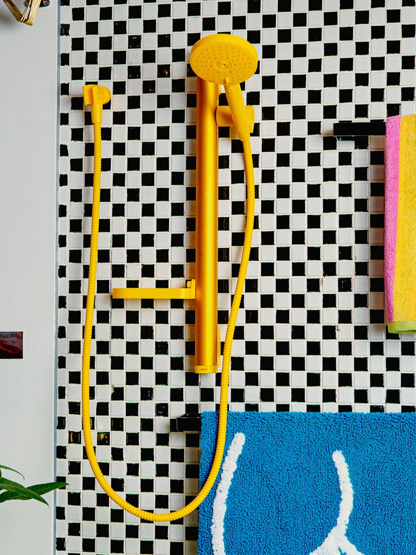 A yellow Minimalist Shower Head installed on a black and white checkered tiled wall besides towel racks.