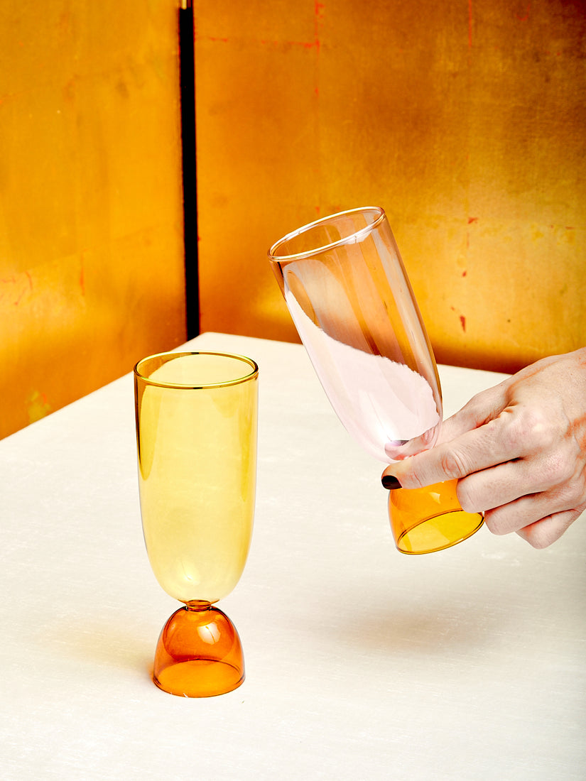 A yellow and amber Harry Highball glass by Mamo sitting next to a pink and amber glass being lifted.