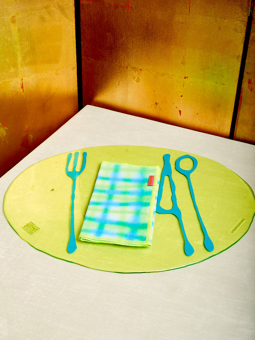 A neon green and blue grid airbrushed napkin is folded showing it's hot pink tag. Underneath is a yellow and turquoise transparent placemat by Gaetano Pesce for Fish Design.