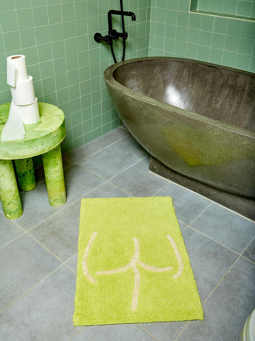 A highlighter green Tushy Bath Mat by Cold Picnic in front of a stone tub and green side table.