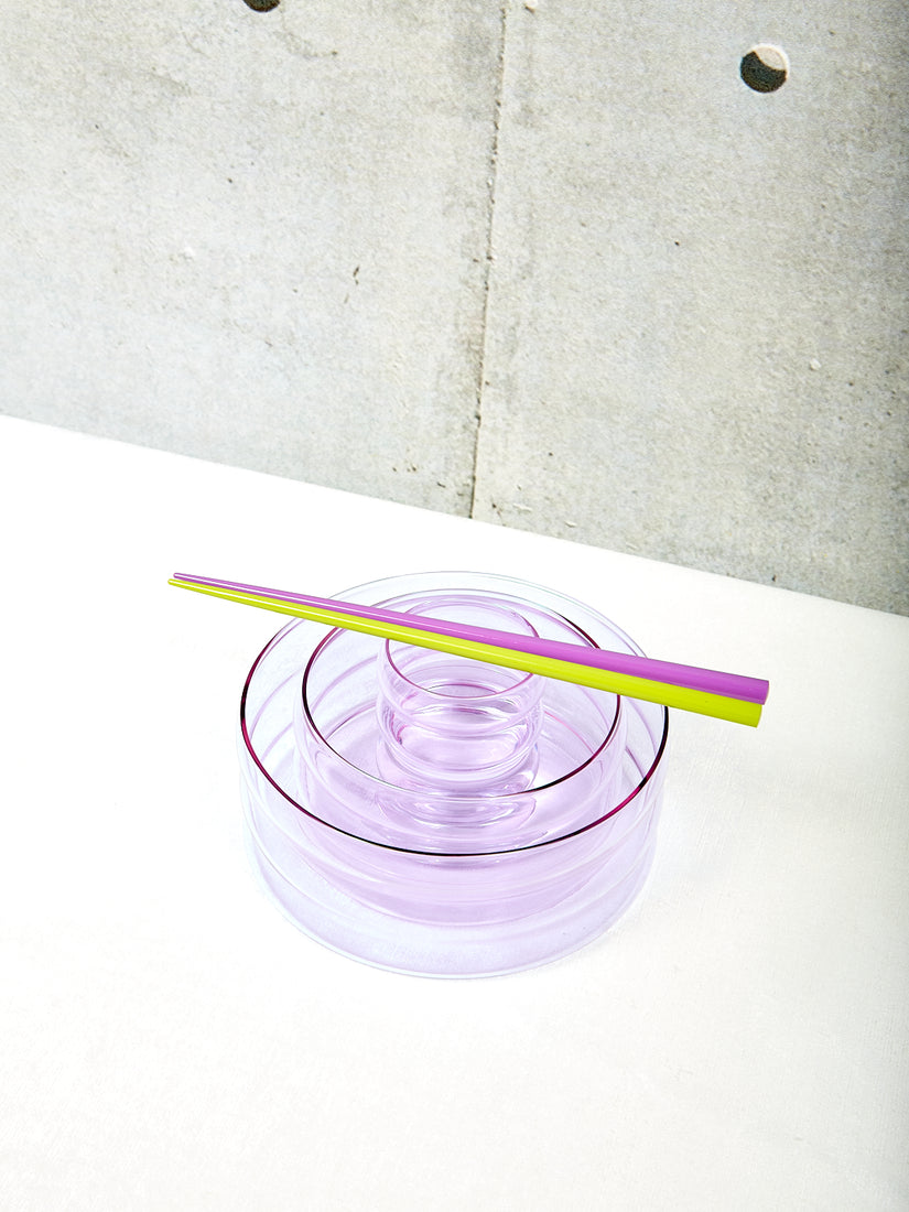 A pair of purple and green chopsticks lays across a nesting of lavender ripple cup, small and medium lavender ripple bowls.