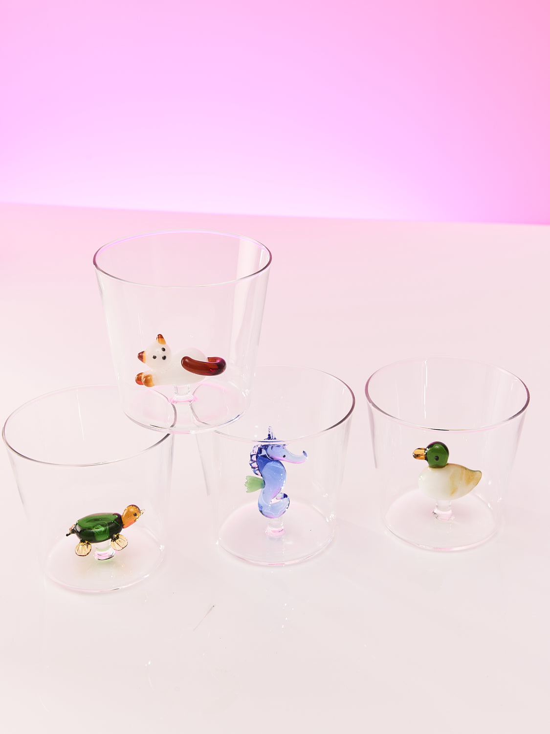 A set of four small cups with glass figurines sit on a cream table with a pink background. The first cup has turtle figure the second cup has a seahorse figure the third cup has a duck figure and the fourth cup has a cat laying down figure.