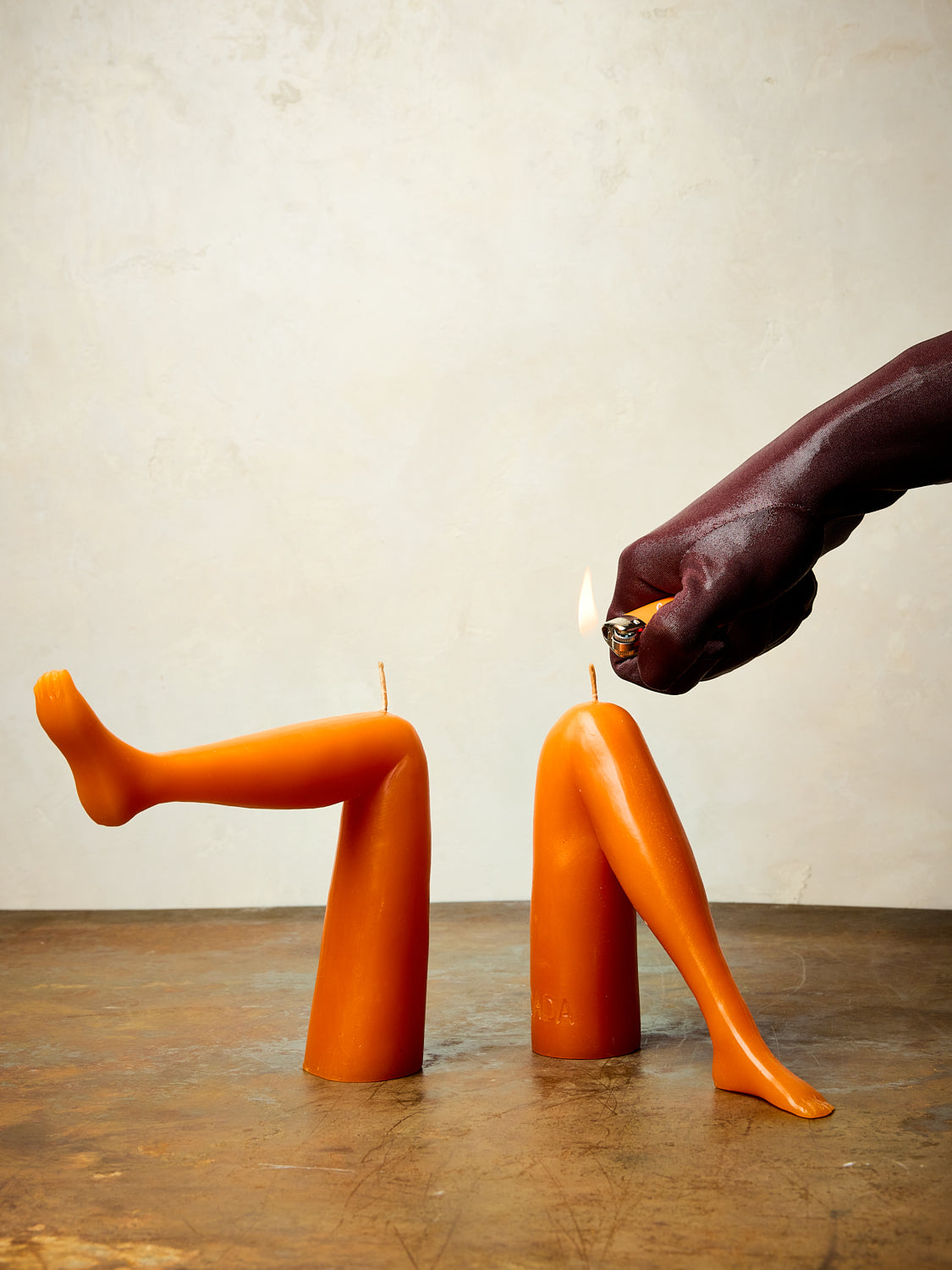 A pair of orange leg candles sit on a table with a persons hand in black glove lighting one of the knees. 