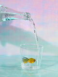 A small cup with a yellow fish made of glass inside it sits on a glass table. Someone is pouring water from a clear bottle into the cup.