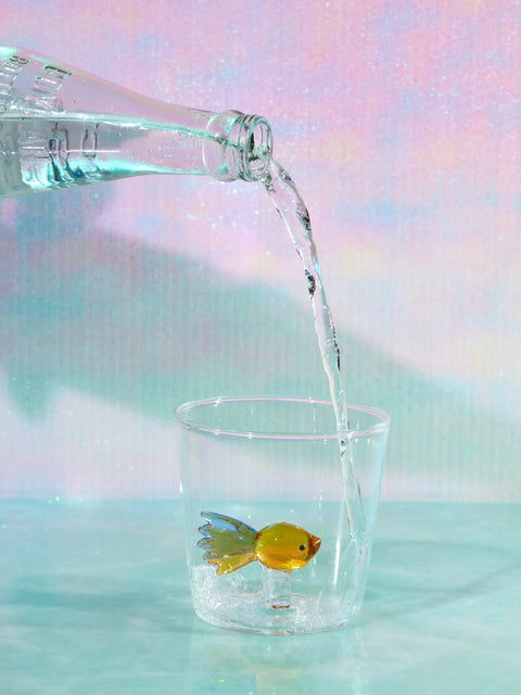 A small cup with a yellow fish made of glass inside it sits on a glass table. Someone is pouring water from a clear bottle into the cup.