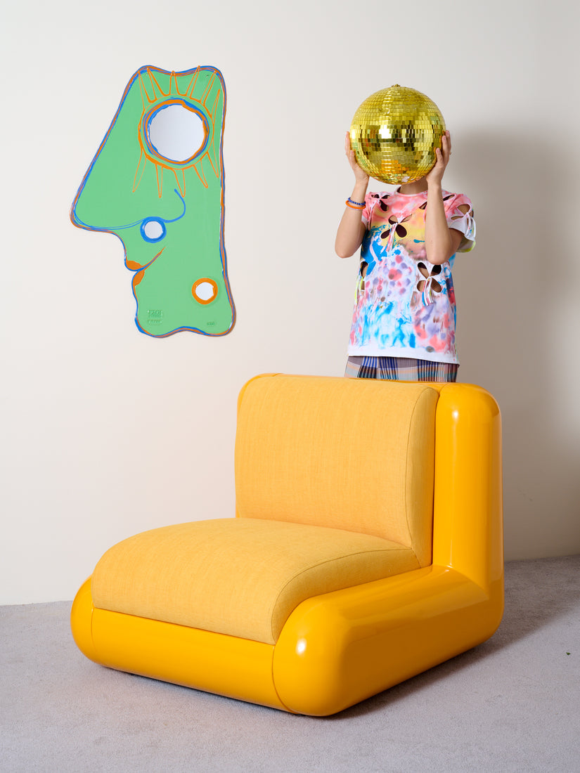 Someone hold a disco ball in front of their face standing behind a yellow Uma T4 chair. On the wall behind the chair is a mint Look At Me Mirror.