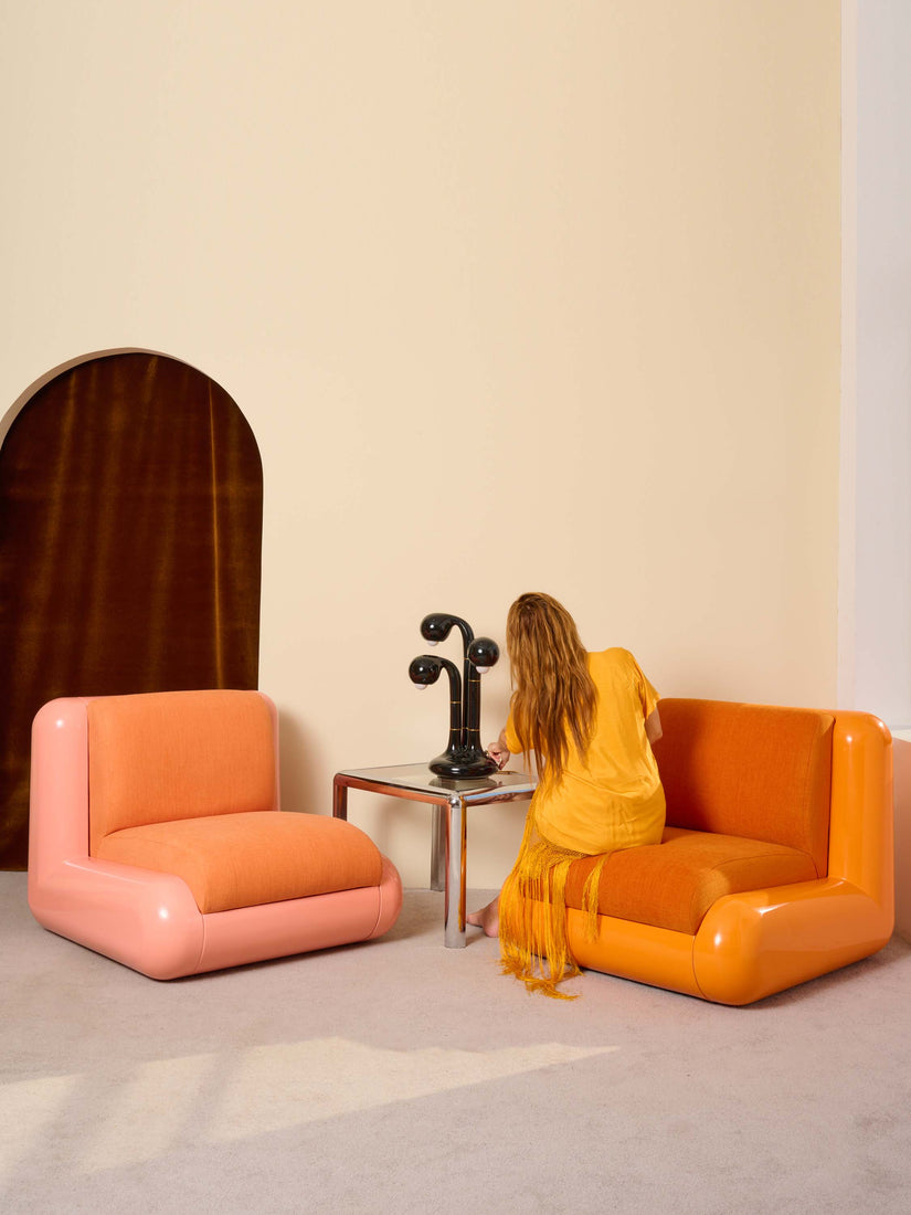 Helena sits on an Orange Uma T4 Chair facing away and switching on a black Entler table lamp. The lamp sits atop a chrome side table between the orange chair and a pink chair.