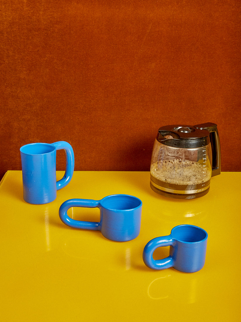 Stoneware Mugs by Workaday Handmade in three sizes sit in a diagonal line next to a pot of coffee.