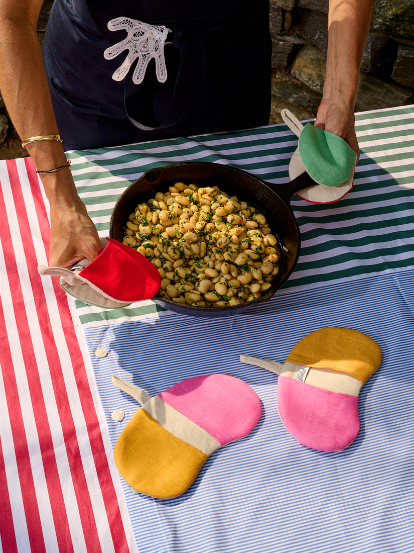 Someone wearing red/mint Bean Pot Mitts by Gohar World handling a cast iron skillet full of beans. They're placing the skillet on a striped table cloth that also has orange/pink Bean Pot Mitts atop.