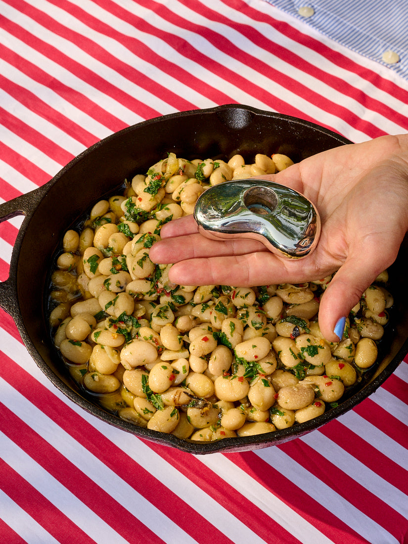A hand holds the sterling silver Bean Candle Holder by Gohar World over a cast iron skillet full of beans.