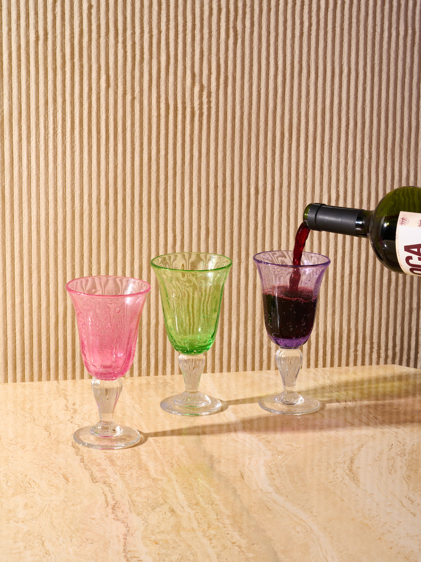 A bottle of red wine is poured into a Purple Bubbled Wine Glass. Sitting left are empty pink and green glasses.