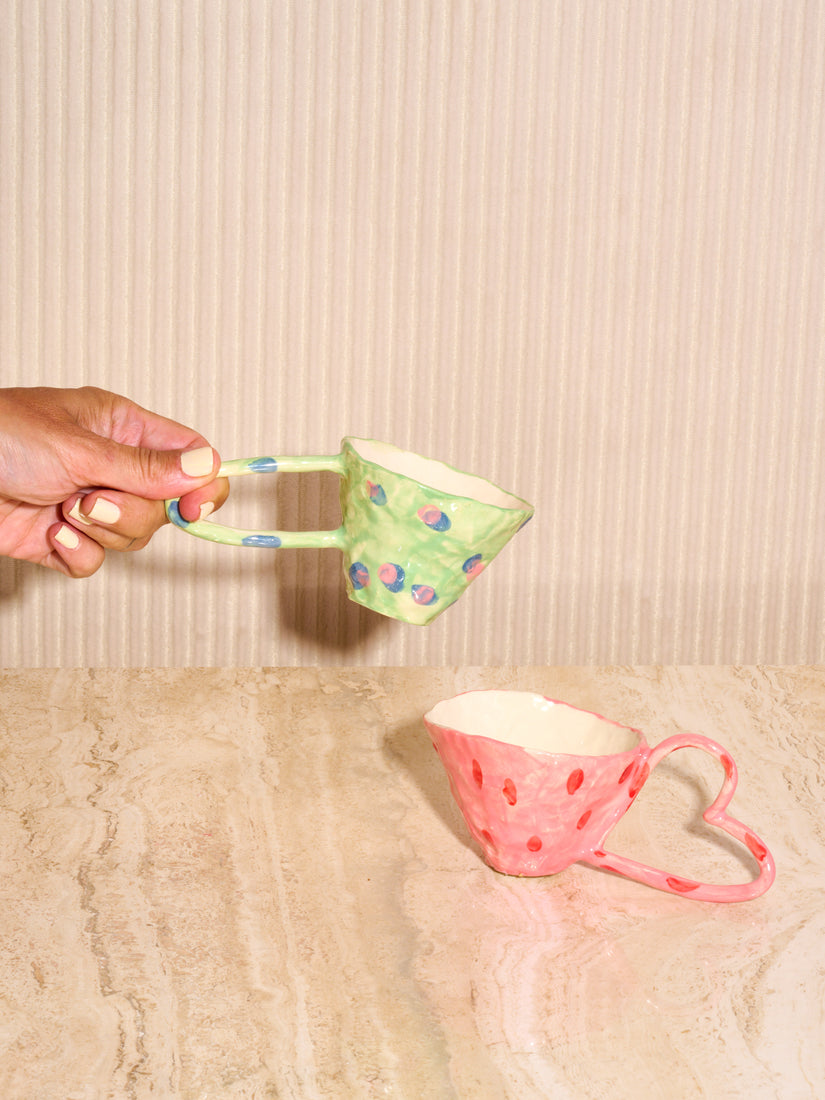 A hand holds up the Green Mug with Peas while the Pink Heart Mug rests below on a travertine table top.