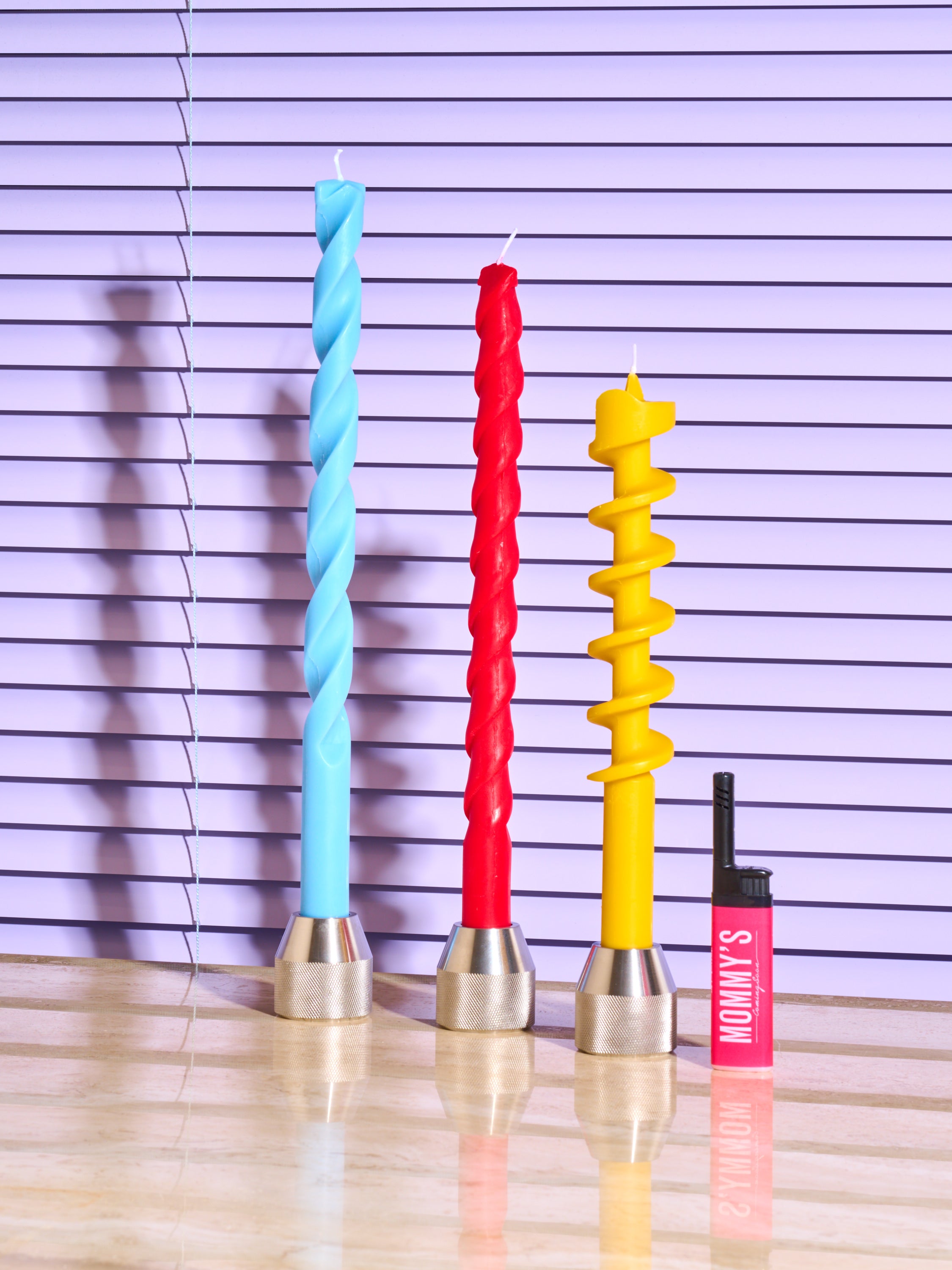 Blue, Red, and Yellow Drill Bit Candles in Drill Bit Candle Holders by 54 Celcius next to a pink Coming Soon lighter.