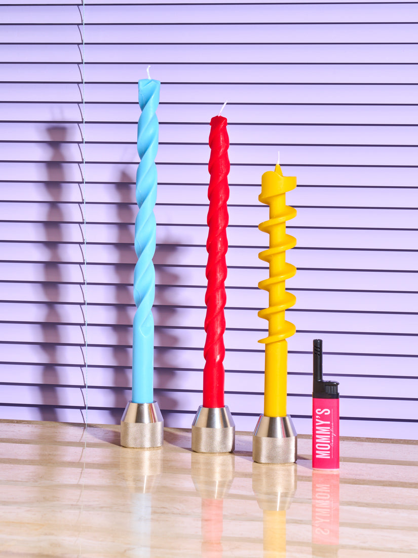 Blue, Red, and Yellow Drill Bit Candles in Drill Bit Candle Holders by 54 Celcius next to a pink Coming Soon lighter.