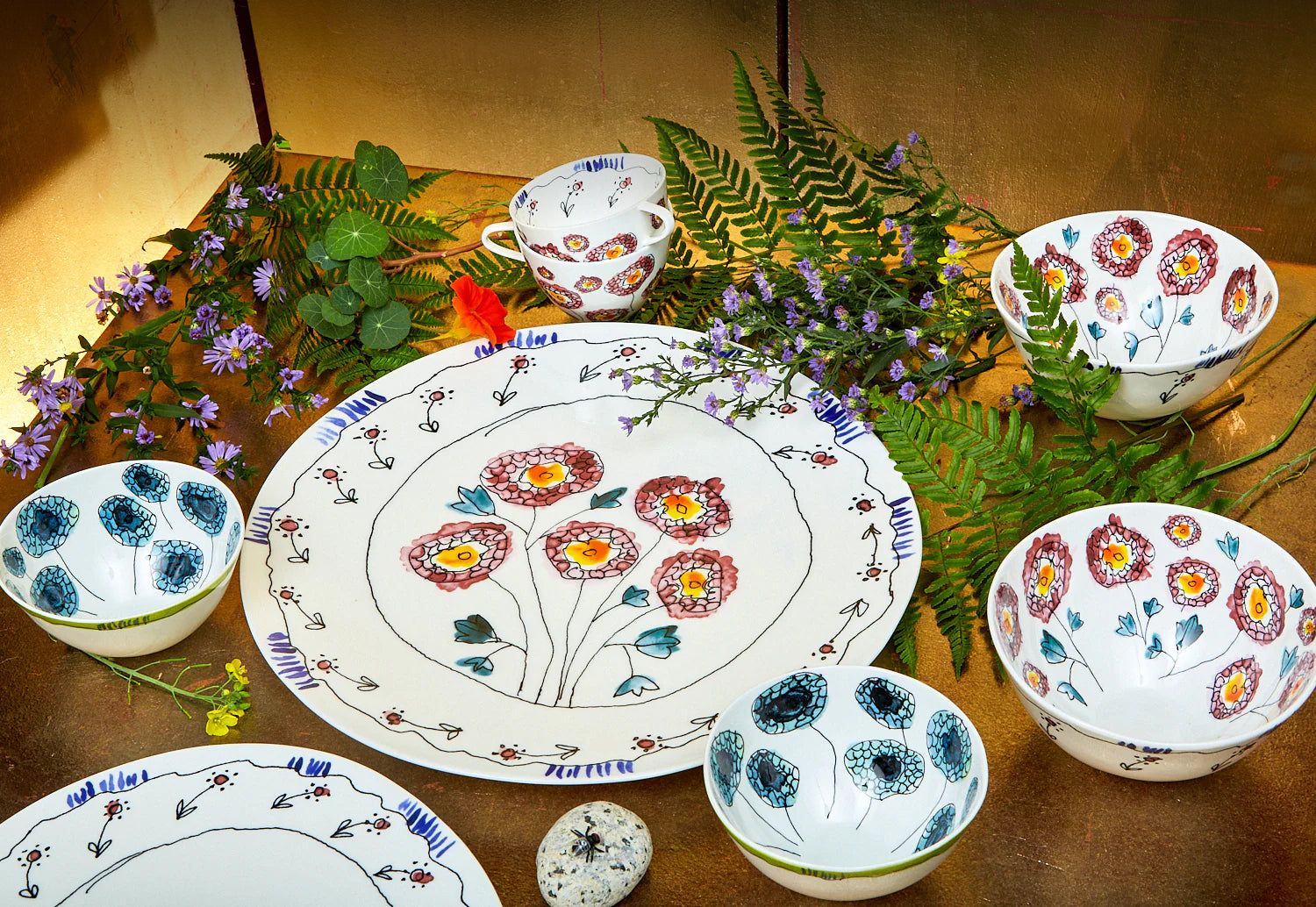 A collection of ceramic plates and bowls with hand drawn flowers on a silver and gold background 