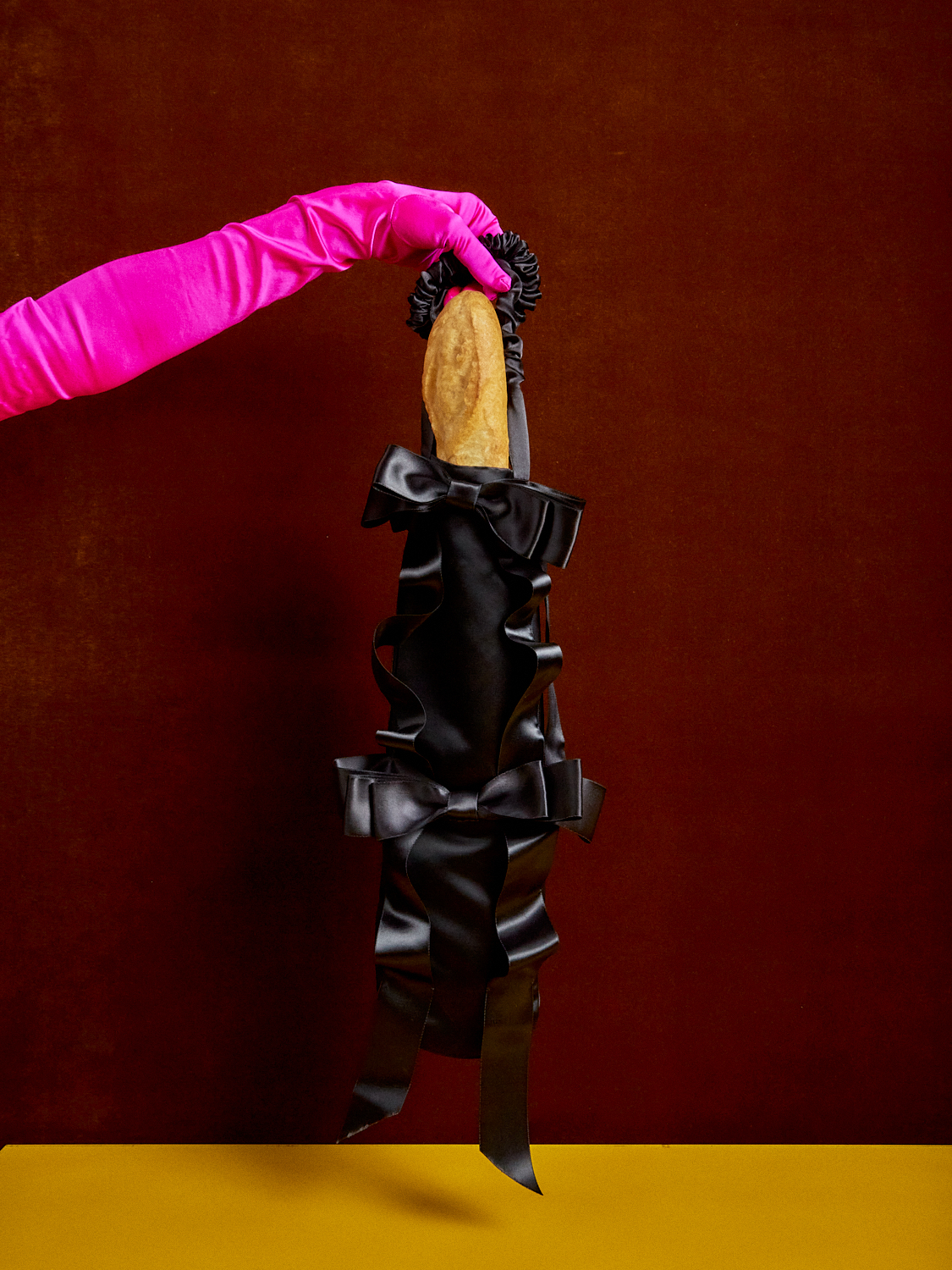 Someone wearing a pink satin glove holds up a black baguette bag full with a baguette.