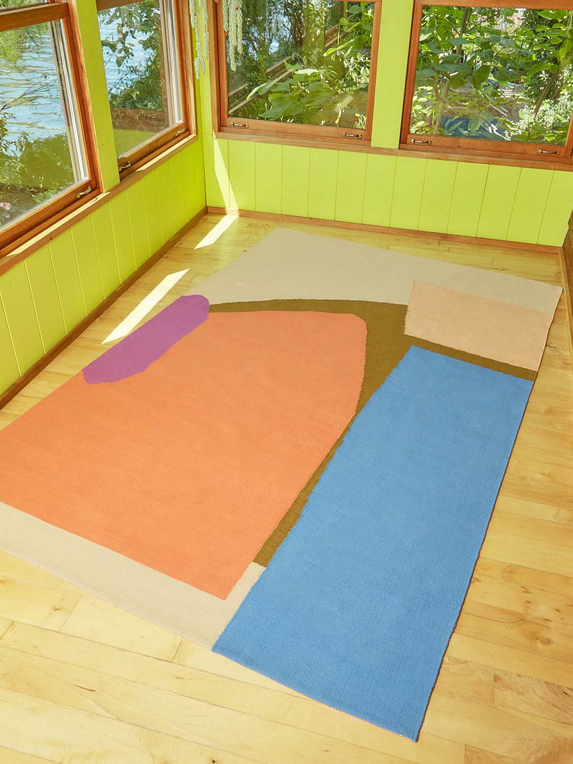 A woven rug by Cold Picnic with orange, blue, magenta, brown, and peach abstract forms laying on hardwood flooring in a lime green room.