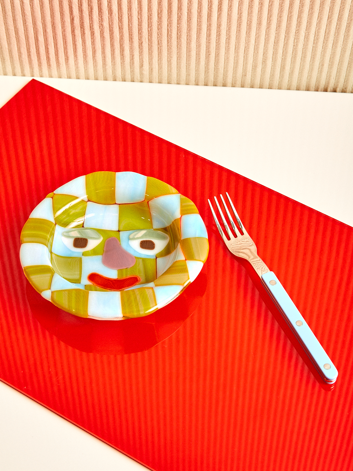 A green and blue checkered glass bowl with a graphic face sits left of a blue Sabre fork.