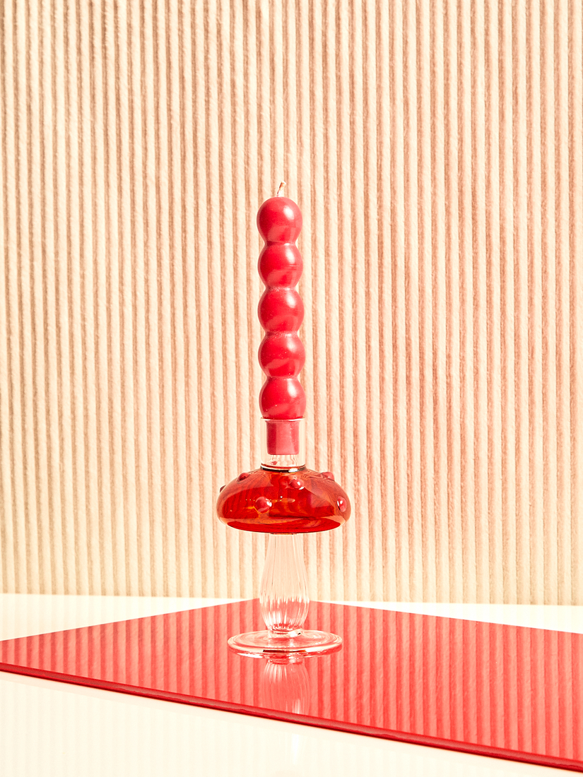 A red Volute Candle by Maison Balzac sits inside of an amber with red dots mushroom candle holder.