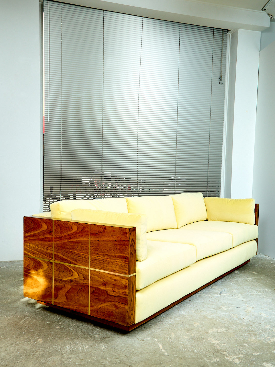 1970s Milo Baughman sofa sits in a room with the shades down in the back. The couch is angled so you can see the side and its wooden panelling. The fabric is velvet with a butter yellow color.